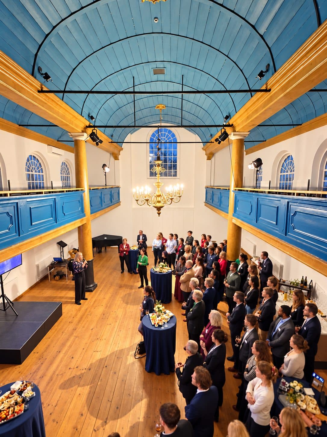 A corporate gathering in a historic hall with a blue vaulted ceiling, a speaker and guests standing